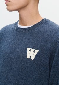 Námornícka modrá pletená sveter s rebrovaným výstrihom, s vyšitým bielym písmenom "W" na ľavej hrudi. Mäkká, textúrovaná látka.