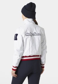 Bílá bombarová bunda s červenými a bílými žebrovanými manžetami a spodním okrajem. Obsahuje vyšitý logotyp "Helly Hansen" a námořnické akcenty na rukávech.