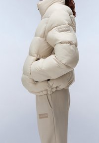 Person i en beige puffjacka och matchande beige byxor, med händerna i jackfickorna, på en enkel ljus bakgrund.