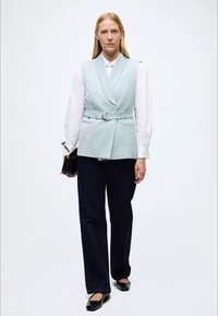 Colete azul claro feito à medida com cinto, usado sobre uma blusa branca, combinado com calças largas azul-escuro e sapatos planos pretos.