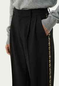 Zwarte op maat gemaakte broek met een hoge taille, voorzien van een gouden decoratieve rand langs de zijzak. Gemaakt van een soepele stof.