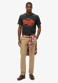 Camiseta de algodón negra con gráfico rojo de "SuperDry", pantalones caqui, camisa de cuadros roja atada a la cintura, zapatos de barco marrones con cordones, look casual.