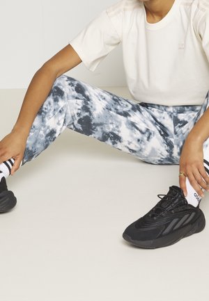 Bílé tričko s jemným logem, modré batikované kalhoty, černé sportovní boty a černobílé pruhované ponožky. Uvolněná sedící póza.