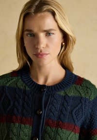 Pletený kardigán s okrúhlym výstrihom. Má viacerými farbami pruhy v námorníckej modrej, zelenej a burgundskej farbe. Tlačidlá sú vyrobené z tmavohnedého materiálu.