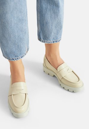 Mocasines de cuero crema con una correa texturizada y una suela de goma gruesa y de color claro, combinados con vaqueros de pierna recta en azul claro.