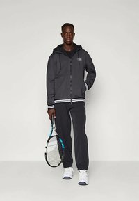 Μαύρο φούτερ με φερμουάρ και λευκές ρίγες, συνδυασμένο με μαύρο παντελόνι, κρατώντας ένα ρακέτα τένις. Περιέχει λογότυπο στο στήθος και αθλητικό σχέδιο.