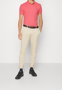 Rotes Poloshirt mit Kragen, beige Chinos, brauer Ledergürtel und schwarze Sneaker. Das Poloshirt hat ein kleines Logo auf der linken Brust.