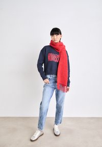 Sudadera marina con texto gráfico rojo, combinada con una larga bufanda roja. Unos vaqueros rectos de color azul claro y unas zapatillas plateadas completan el atuendo.