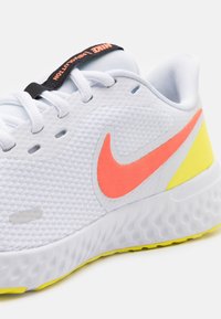Běžecká obuv Nike s bílým síťovaným svrškem, neonově růžovým logem Swoosh, žlutou patou, texturovanou podešví a černou poutkem pro snadné obutí.