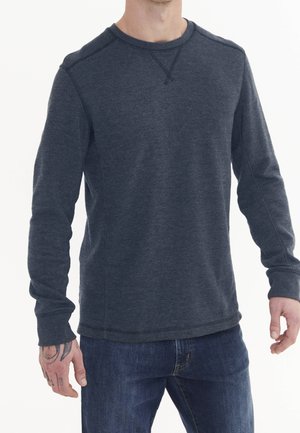 Mężczyzna ubrany w ciemnoszarą teksturowaną koszulę z długim rękawem i niebieskie dżinsy, z widocznymi tatuażami na lewej ręce, stojący na białym tle.