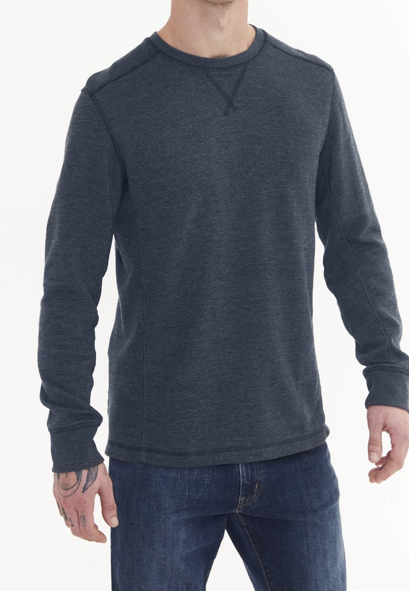 Homme portant une chemise texturée à manches longues gris foncé et un jean bleu, avec des tatouages visibles sur sa main gauche, debout devant un fond blanc.