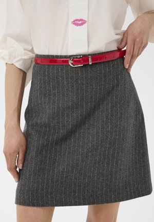 Chemise blanche avec trace de rouge à lèvres rose associée à une jupe grise à fines rayures et une ceinture rouge brillante attachée à la taille.