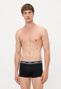 Calzoncillos boxer negros con una cinturilla con logo en gris, hechos de material elástico. Modelo con el cabello corto posando de pie contra un fondo sencillo.