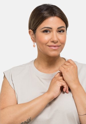 Boucles d'oreilles en or rose avec un design en éclat de soleil, portées par une personne dans un top sans manches gris clair, bras croisés au premier plan.