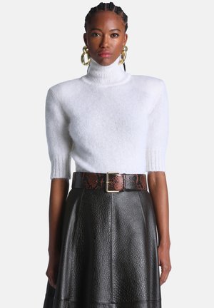 Femme portant un pull à col roulé en maille blanche avec des manches jusqu'aux coudes et une jupe en cuir noir texturé avec une large ceinture marron.