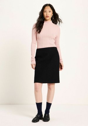 Frau mit langen dunklen Haaren, die einen rosa gerippten Rollkragenpullover, einen schwarzen knielangen Rock, marineblaue Socken und schwarze Loafers trägt und vor weißem Hintergrund steht.