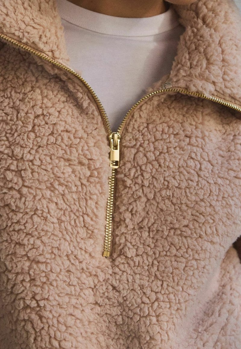 Løs, lyserød fleecejakke med lynlås i guld, der har en blød tekstur og et minimalistisk design. Hvid skjorte synlig under.