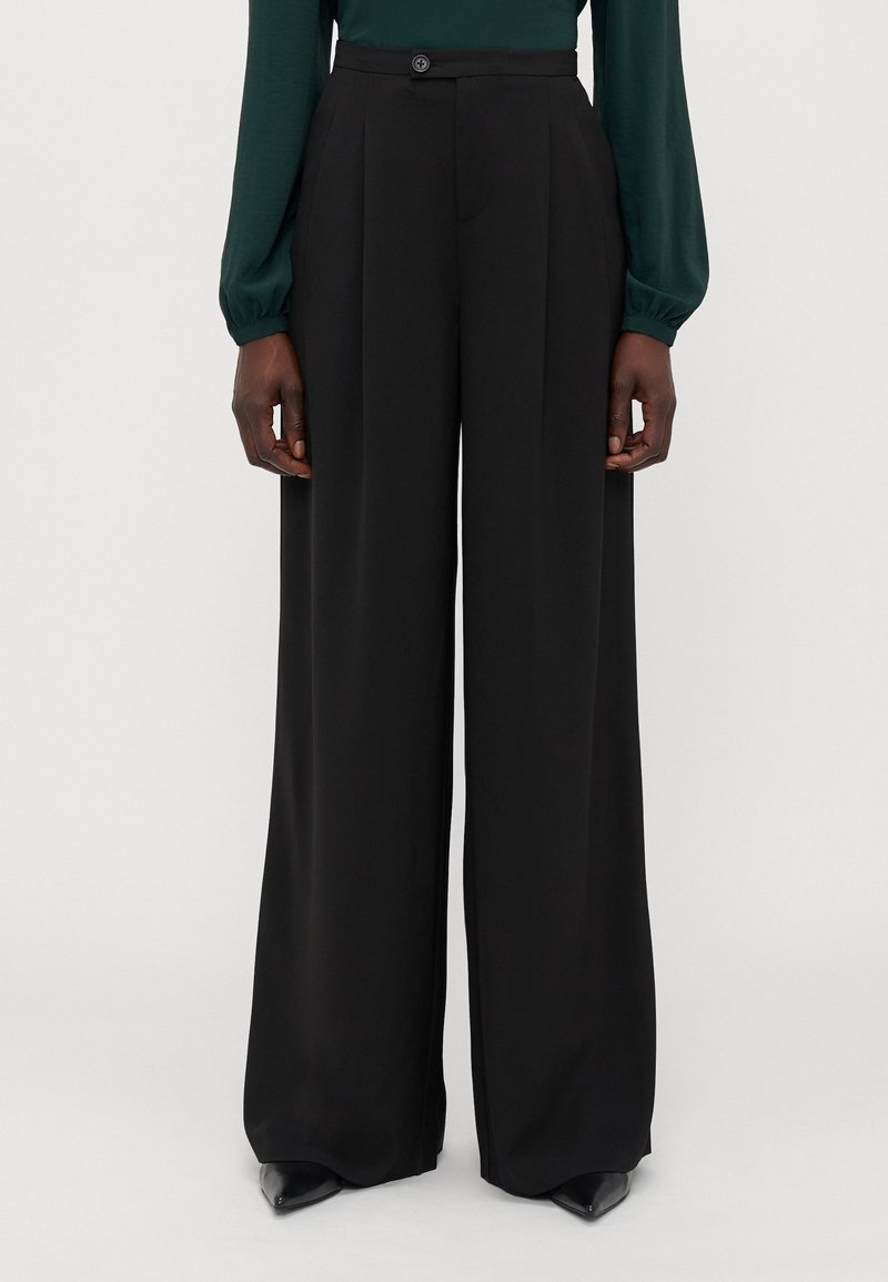 Pantalon noir taille haute à jambes larges avec plis devant, porté avec un haut manches longues vert foncé et des chaussures noires pointues.