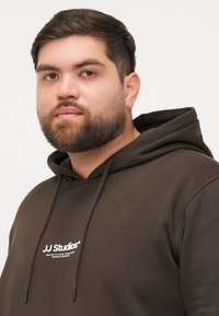 Haut à capuche marron avec un cordon de serrage, arborant un logo blanc "JJ Studios" sur la poitrine. Tissu doux avec une coupe standard.