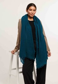 Écharpe tissée turquoise à bords effilochés, superposée sur un haut noir et un blazer beige, le modèle pose avec une main sur un tabouret en bois blanc.