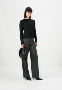 Donna che indossa un maglione nero a coste, pantaloni grigi a gamba larga, décolleté neri e tiene una piccola borsa in pelle nera.