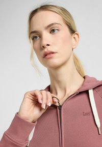 Sudadera con cremallera de color rosa con cordones blancos, puños acanalados y textura suave; presenta un pequeño logo bordado en el pecho.