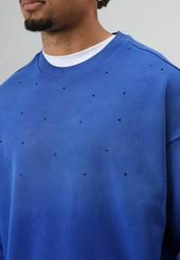 Sweatshirt bleu en tissu doux, avec un col rond et de petites décorations noires dispersées sur le devant.