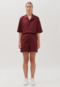 Camicia a maniche corte con bottoni e pantaloni corti abbinati in un profondo bordeaux, realizzati in tessuto liscio, con tasca sulla camicia e vita elastica.
