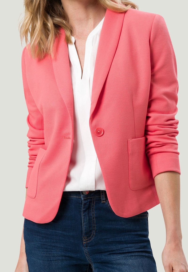 Femme portant un blazer corail ajusté sur une blouse blanche et un jean bleu foncé, avec les manches remontées jusqu'aux avant-bras.