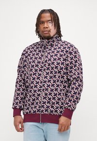 Veste zippée en marine, bordeaux et blanc avec un motif géométrique. Présente un col haut et des poignets côtelés. Texture de tissu douce.