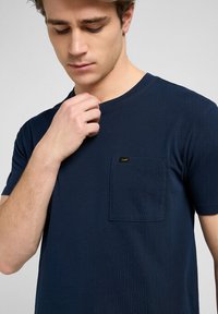 Granatowa koszulka z krótkim rękawem z miękkiego materiału, z okrągłym dekoltem, z kieszonką na lewej piersi oraz subtelną metką marki. Ogólnie gładka tekstura.