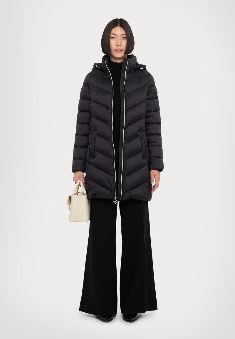 Doudoune noire avec un motif matelassé horizontal, fermeture éclair dorée et capuche. Le mannequin tient un petit sac à main crème, portant un pantalon noir large.