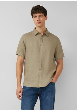 Mann mit dunklem lockigem Haar trägt ein beiges kurzärmeliges Hemd und blaue Jeans, steht mit einer Hand in der Tasche.