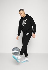 Zwarte Nike hoodie met wit logo, bijpassende zwarte sweatpants en witte schoenen met blauwe accenten. Een voetbal ligt dichtbij de voet.
