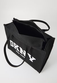 Juodas kanvaso tote krepšys su viršutiniu užtrauktuku, dviem tvirtais rankenomis ir baltu spausdintu "DKNY NEW YORK" logotipu priekyje. Vidus nėra išklotas.