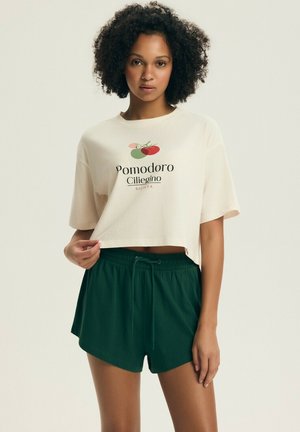 Kobieta z kręconymi włosami w beżowym, krótkim t-shircie z grafiką pomidora i napisem, w ciemnozielonych szortach ze sznurkiem, stoi na jednolitym tle.