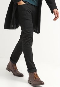 Černé slim-fit kalhoty v kombinaci s hnědými kotníkovými botami s oranžovými tkaničkami. Oblečení doplňuje dlouhý černý kabát a modrá košile.