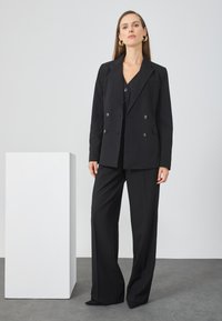 Černý oblek na míru s dvouřadým sakem se lesklými knoflíky a kalhotami širokého střihu, umístěný na neutrálním pozadí.