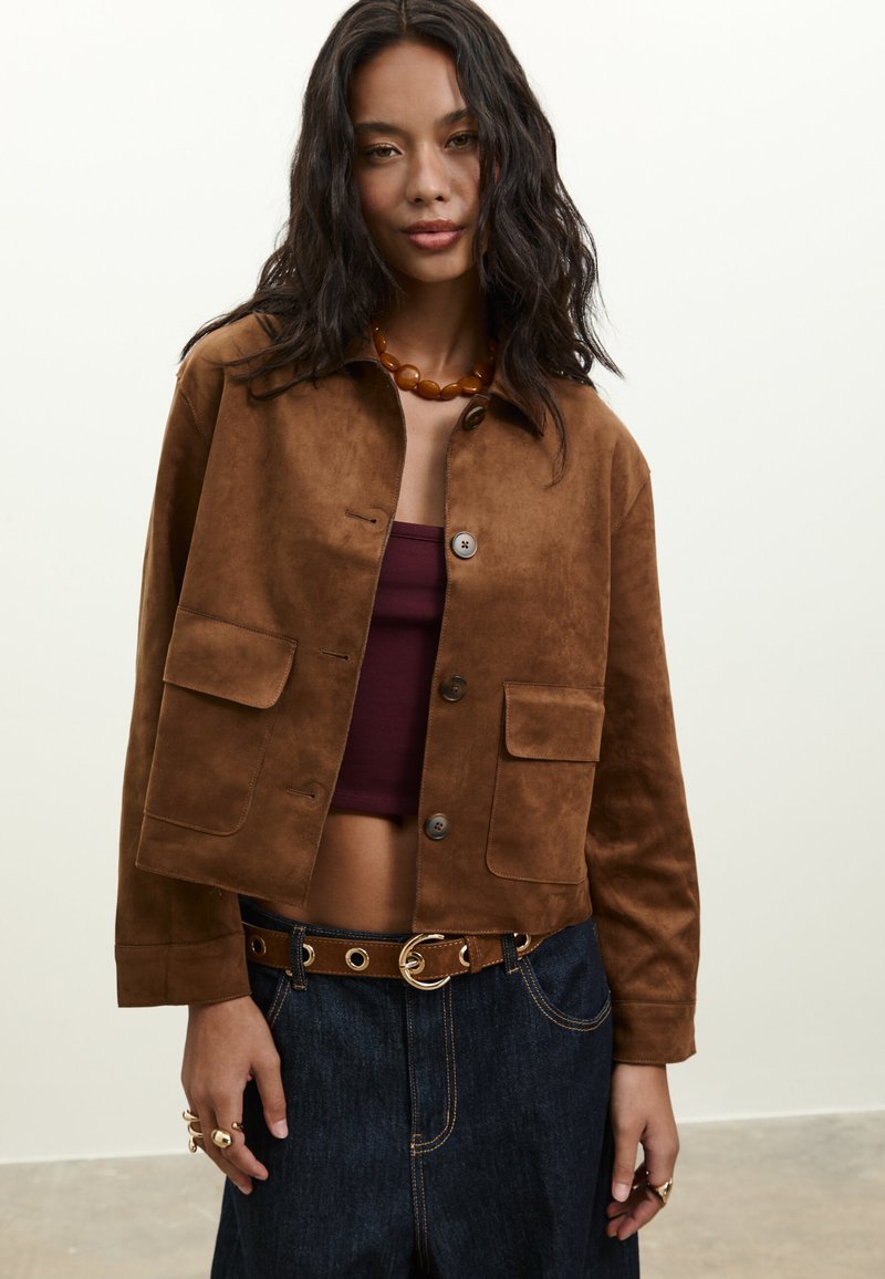 Frau trägt eine braune Wildlederjacke über einem dunkelburgunderfarbenen Trägertop, dunkle Jeans mit braunem Gürtel, bernsteinfarbenen Perlenhalskette und goldene Ringe.