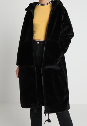 Person iført en lang sort hætteklædt faux pelsjakke over en sennepsgul croppet ribbet trøje og sorte bukser med høj talje.