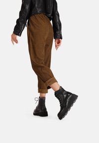 Stivali neri in pelle fino alla caviglia con suola spessa, lacci e cuciture decorative. Abbinati a pantaloni in velluto a coste marroni arrotolati.