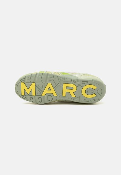 Semelle d’une basket vert clair avec de grandes lettres jaunes écrivant MARC sur le dessous, vue de dessous sur fond blanc.