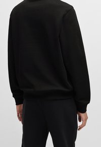 Felpa nera con collo rotondo e maniche lunghe, caratterizzata da una texture liscia e polsini a coste. Nessun logo o motivo visibile.