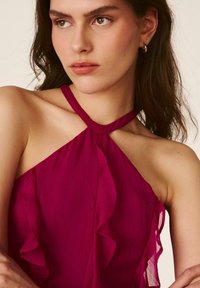 Joven con cabello castaño medio que lleva una camiseta sin mangas de color magenta oscuro con detalles de volantes, mirando ligeramente hacia un lado.