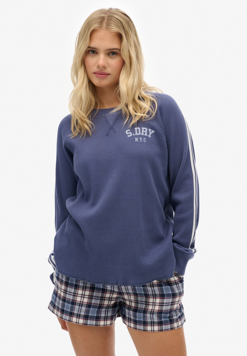 Sudadera de punto azul con puños acanalados y logo blanco. Combinada con pantalones cortos de cuadros en colores azul, blanco y burdeos.