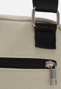 Beige textured synthetic bag featuring a contrasting black strap with metal accents and a zippered closure on the front panel.