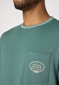 Maglietta in cotone verde con colletto tondo, bordo a righe a contrasto e una tasca frontale con un logo in bianco e dettagli floreali.