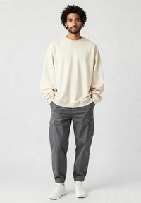 Felpa oversize beige, pantaloni cargo grigi con polsini elasticizzati e sneakers bianche. Outfit casual con dettagli a coste sui polsini.