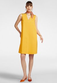 Abito giallo senza maniche con scollo a V, bottoni decorativi sulle spalle e silhouette dritta. Abbinato a tacchi arancioni a punta.