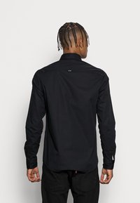 Camicia nera a maniche lunghe con colletto alla coreana, texture liscia e design aderente. Presenta un logo discreto sulla schiena e polsini con bottoni.